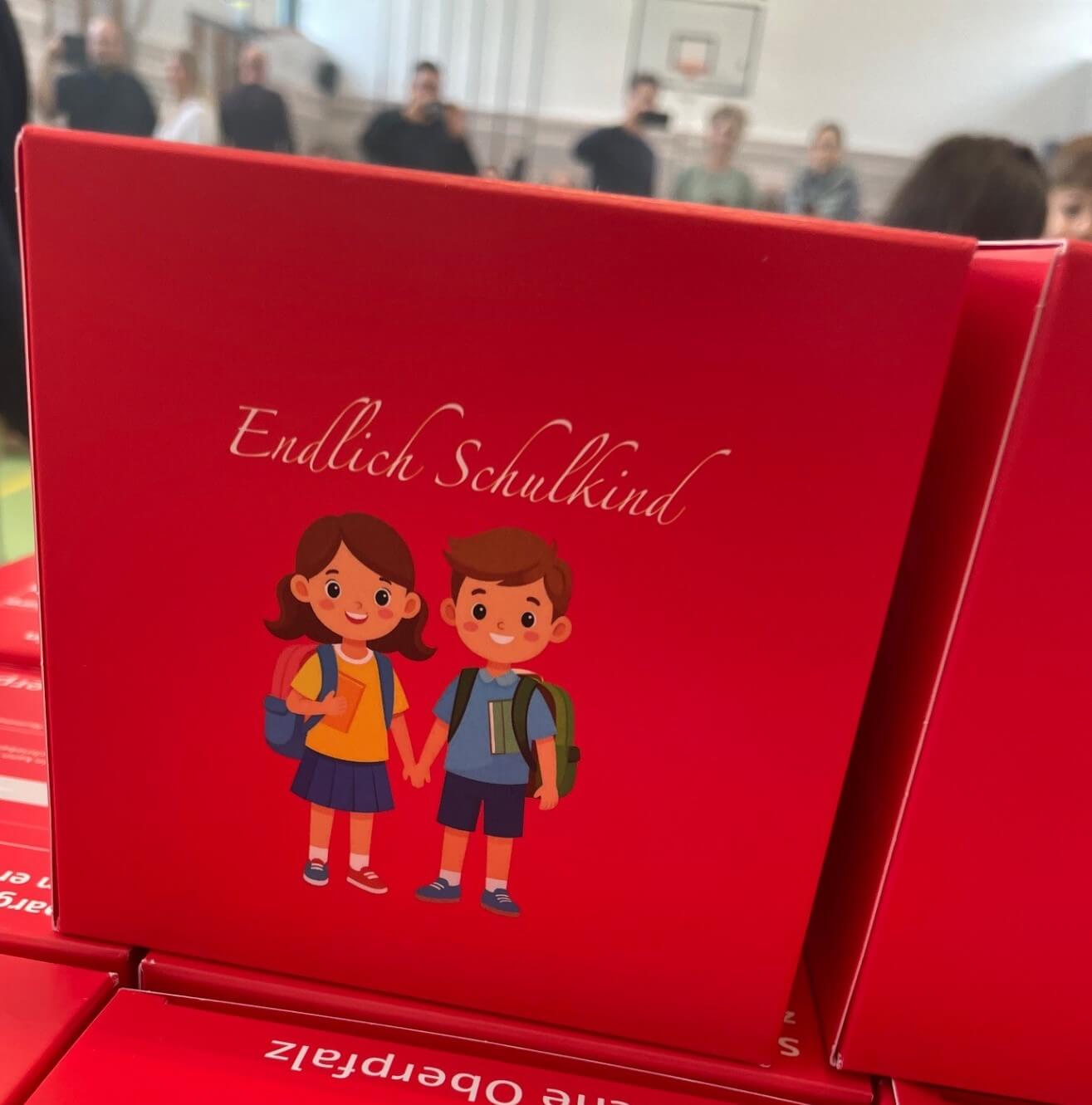 roter Karton mit weißem Schriftzug endlich Schulkind, zwei lächelnde Kinder aufgemalt